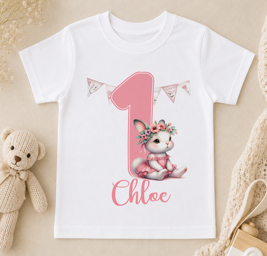 White t-shirt printed with a pink bunny in a flower crown, a pink number one, bunting and a personalised name.