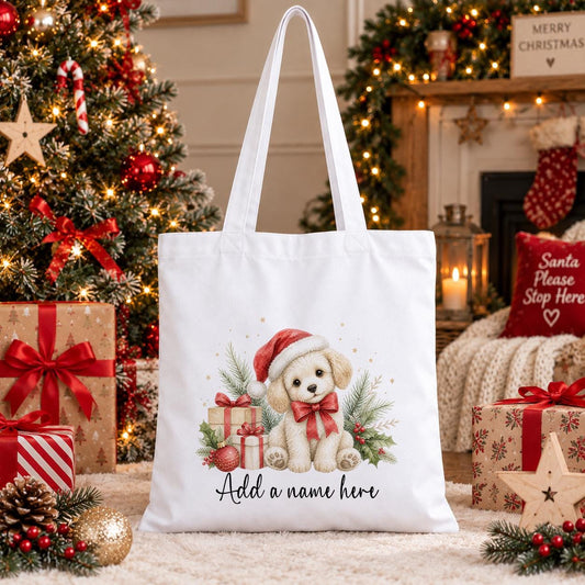 White cotton tote bag printed with a cream puppy in a red Santa hat sitting beside wrapped gifts, baubles and holly, above a name in script.