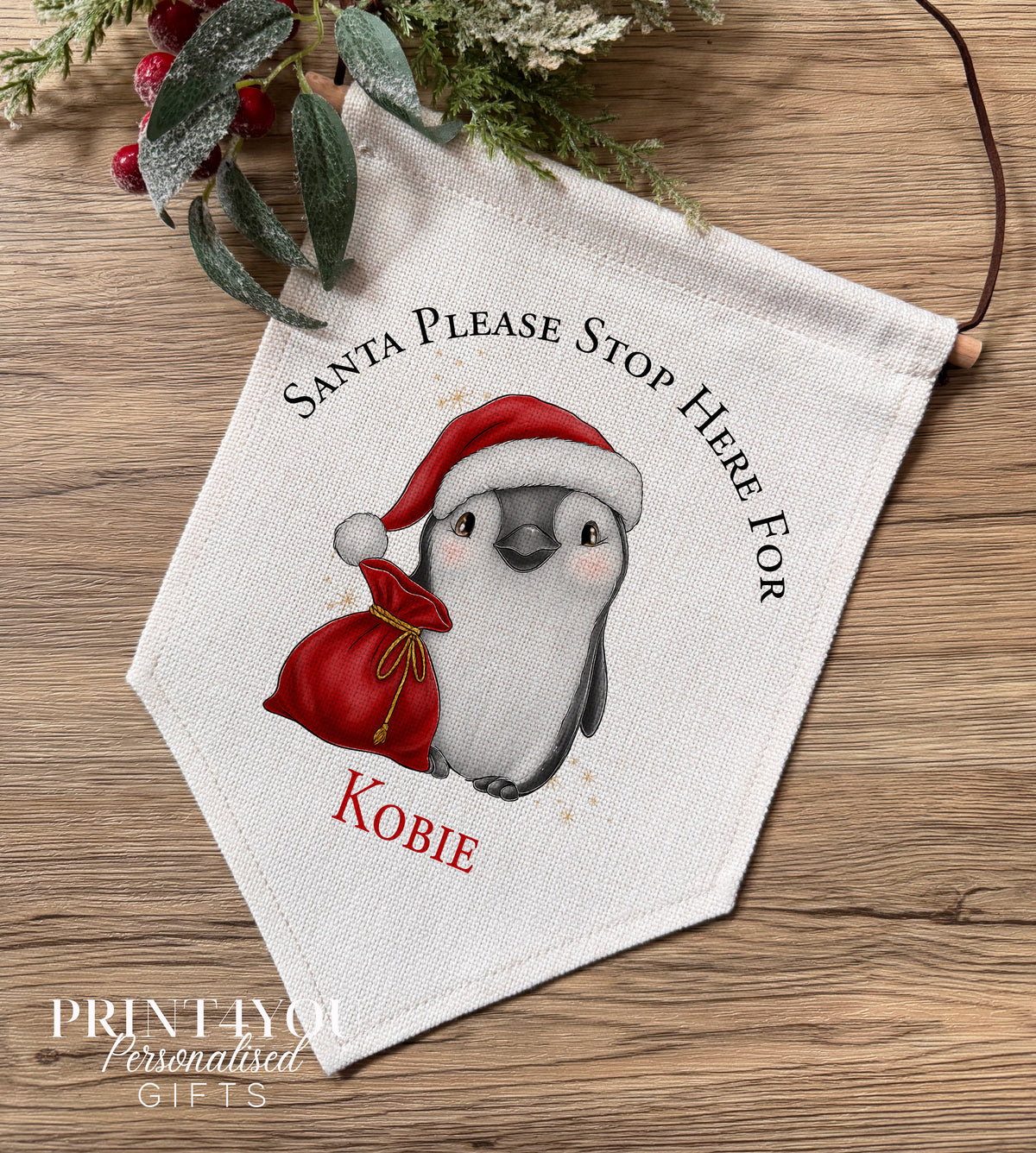 Personalised linen Christmas banner featuring a penguin wearing a Santa hat beside a Santa Stop Here message, personalised with the name Millie, hung on a wooden bar