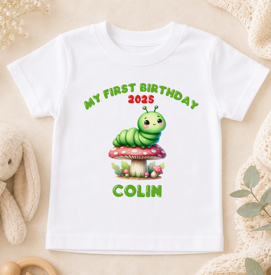 White t-shirt printed with a green caterpillar sitting on a red-spotted toadstool, personalised with 'My First Birthday' and the sample name 'Colin'.