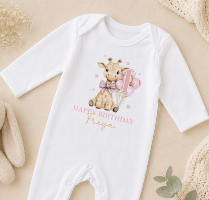 A white onesie with a giraffe and balloons, along with the text "Happy Birthday Freya" printed on it, is displayed on a beige surface.