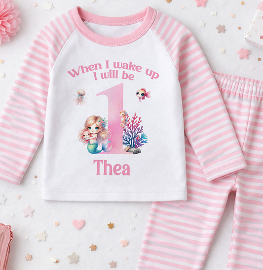 A pink and white striped onesie with a mermaid design and the text "When I wake up I will be Thea" printed on it, along with a small mermaid figure.