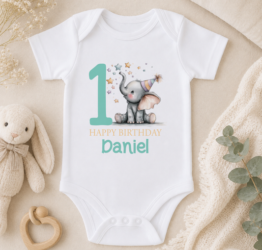 Personalised white baby vest printed with a party-hatted elephant, stars and 'Happy Birthday Daniel', styled on a beige backdrop with a soft toy, a wooden rattle and greenery.