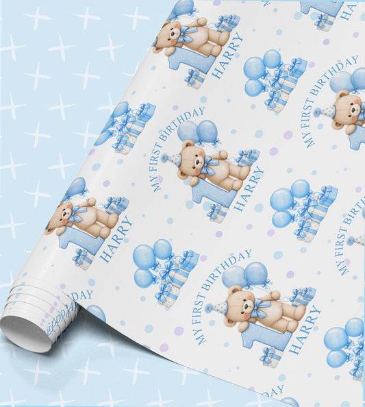 Personalised first birthday wrapping paper with a teddy bear in a blue party hat leaning on a big blue number one, blue balloons and striped gift boxes, printed with My First Birthday and the name Harry on a pale blue sparkle background