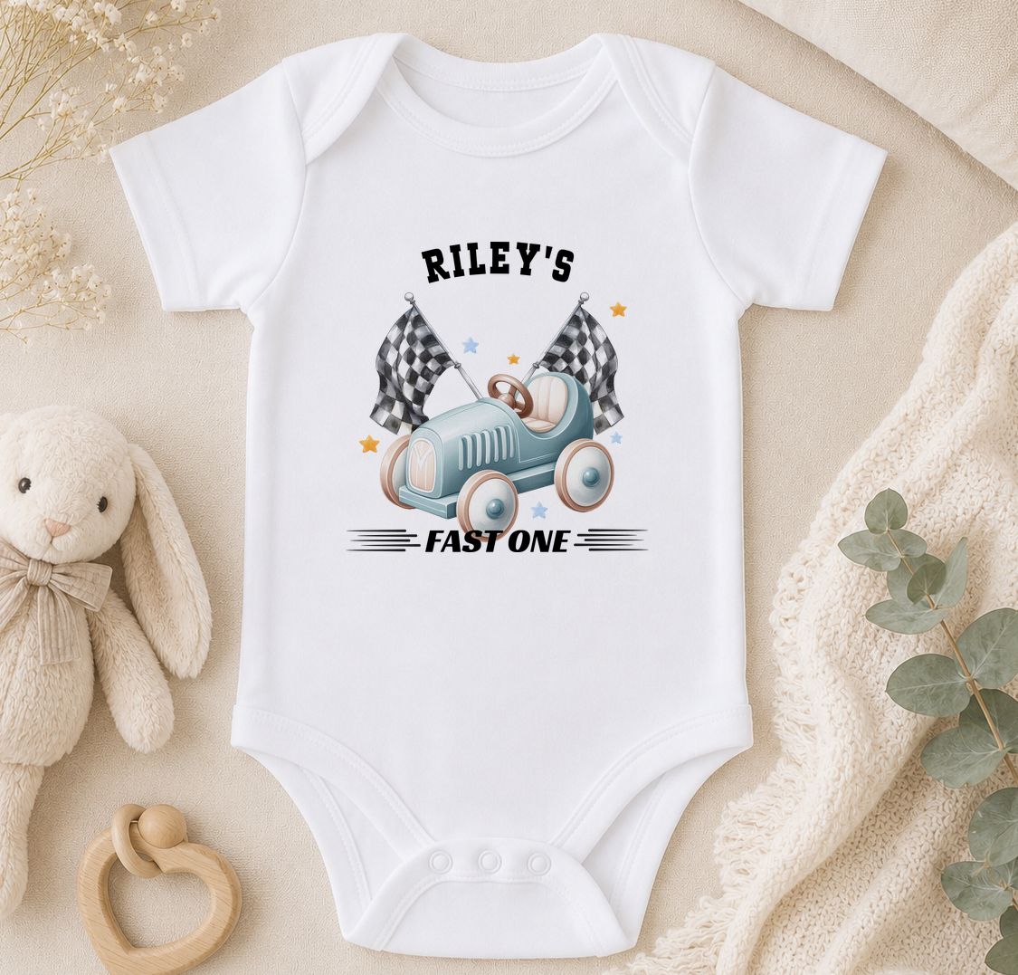A white baby onesie with a graphic of a car and checkered flags, along with the text "Riley's FAST ONE". The onesie is placed on a beige surface, surrounded by a stuffed animal, a baby rattle, and some greenery.