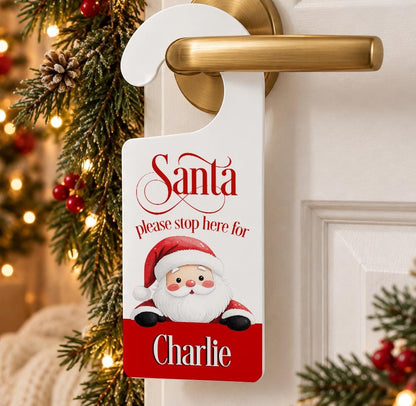 Personalised Santa please stop here door hanger with Santa peeking over a red banner printed with a child’s name, on a door handle
