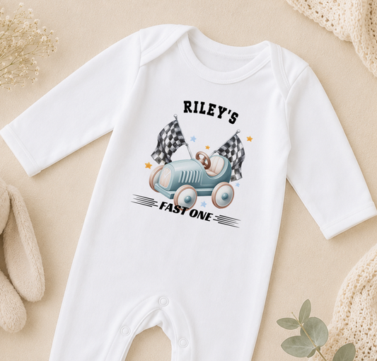 White baby onesie printed with a race car design and the text 'Riley's FAST ONE'.