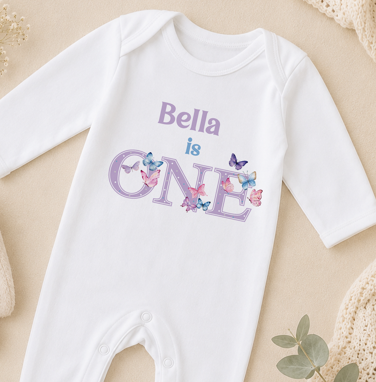 A white onesie with the text "Bella is ONE" printed on it, surrounded by colorful butterflies.