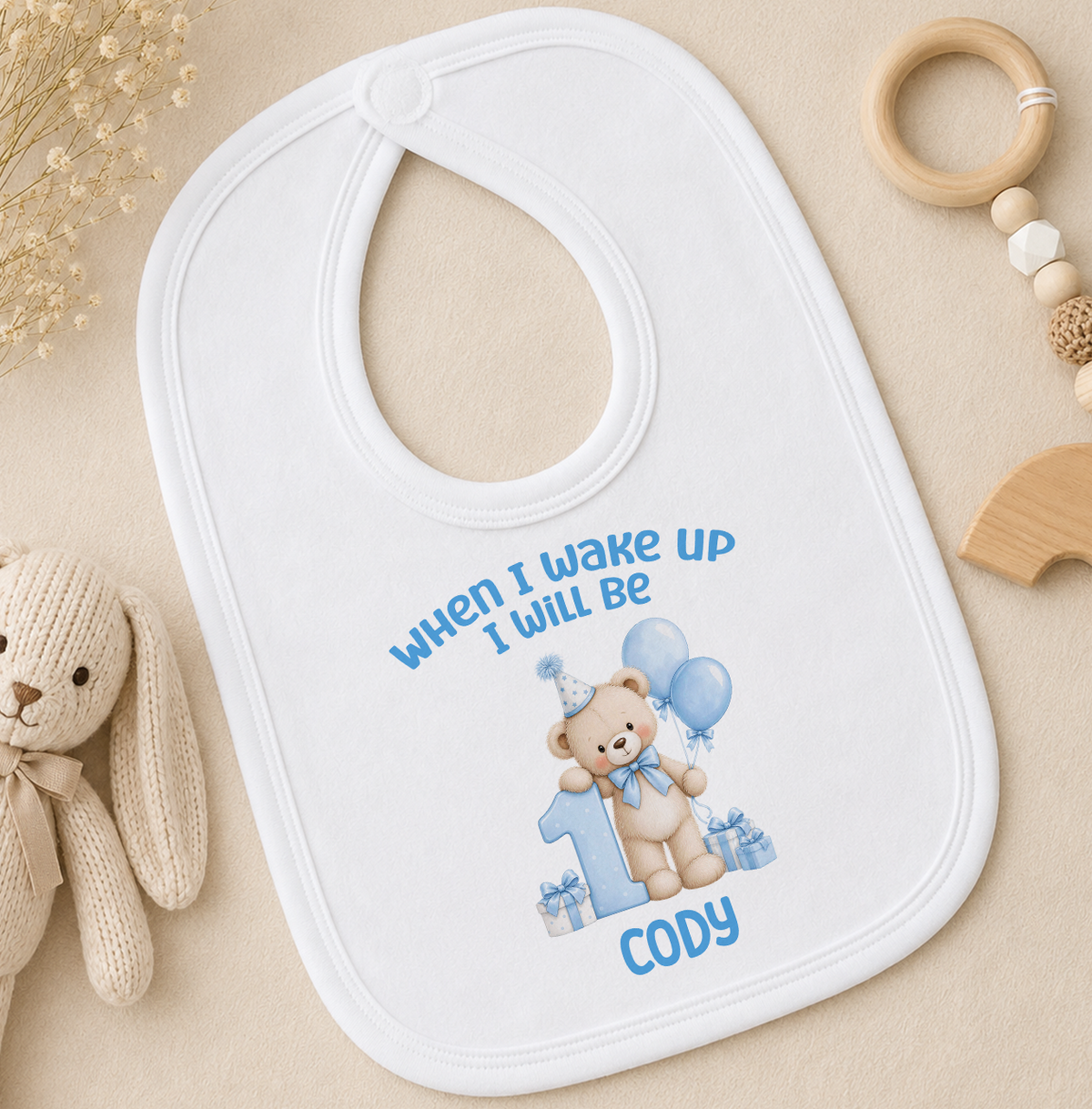 Matching personalised white baby bib with the When I Wake Up I Will Be 1 blue teddy bear and balloons design and the name Cody