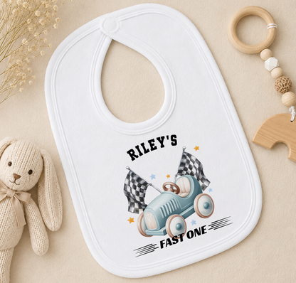 A white baby bib with a personalized design, featuring a race car and checkered flags, is placed on a surface alongside a stuffed animal and a wooden toy ring.