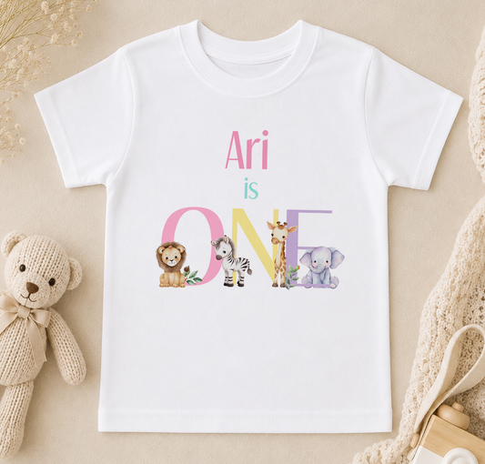 White children's t-shirt printed with 'Ari is ONE' and rainbow safari animal illustrations including a lion, zebra, giraffe and elephant.