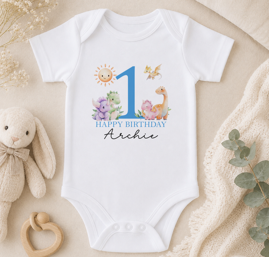A white onesie with a blue number 1 and a "Happy Birthday Archie" message, along with illustrations of various animals, is displayed on a beige surface.