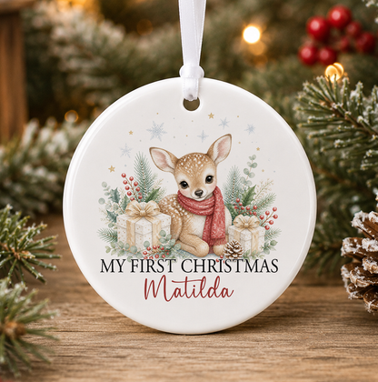 Personalised fawn My First Christmas bauble, white ceramic ornament with a spotted baby deer in a red scarf among cream and gold presents, berries and snowflakes, printed with a baby's name in red script