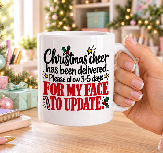 White 11oz ceramic Christmas mug printed with “Christmas cheer has been delivered. Please allow 3–5 days for my face to update.” in black and red lettering with holly, a candy cane and gold stars, held in a festive room