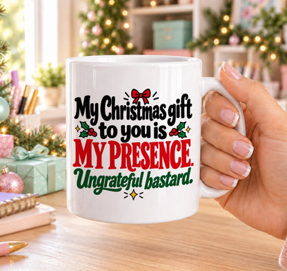 White 11oz ceramic Christmas mug printed with “My Christmas gift to you is my presence. Ungrateful bastard.” in red, green and black lettering with a red bow and holly, held in a festive room