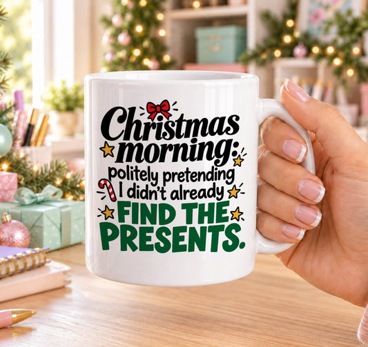 White 11oz ceramic Christmas mug printed with “Christmas morning: politely pretending I didn’t already find the presents.” in black and green lettering with a red bow, candy cane and gold stars, held in a festive room