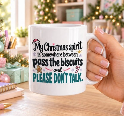 White 11oz ceramic Christmas mug printed with “My Christmas spirit is somewhere between pass the biscuits and please don’t talk.” in black and teal lettering with a Santa hat, gingerbread man, candy cane and snowflakes, held in a festive room