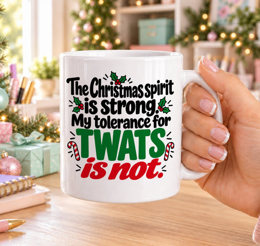 White 11oz ceramic Christmas mug printed with “The Christmas spirit is strong. My tolerance for twats is not.” in red, green and black lettering with candy canes and holly, held in a festive room