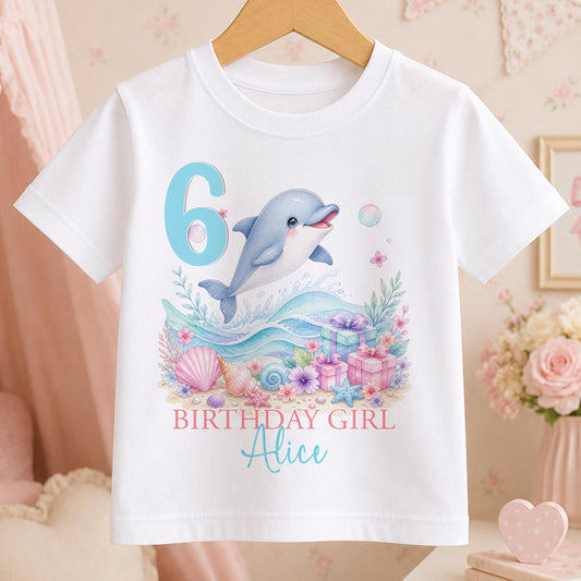White cotton t-shirt printed with a watercolour dolphin leaping over pastel waves beside a large age number, with Birthday Girl and a name below.