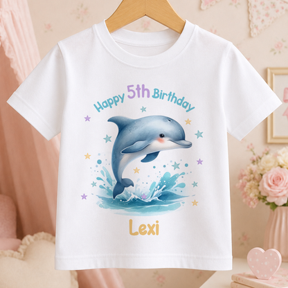 A white t-shirt with a cartoon image of a dolphin jumping out of the water, surrounded by stars and a pink background. The text on the t-shirt reads "Happy 5th Birthday Lexi".