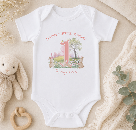 A white onesie with a pink number 1 and a bunny illustration, along with a stuffed bunny and a beige blanket, are arranged on a surface.
