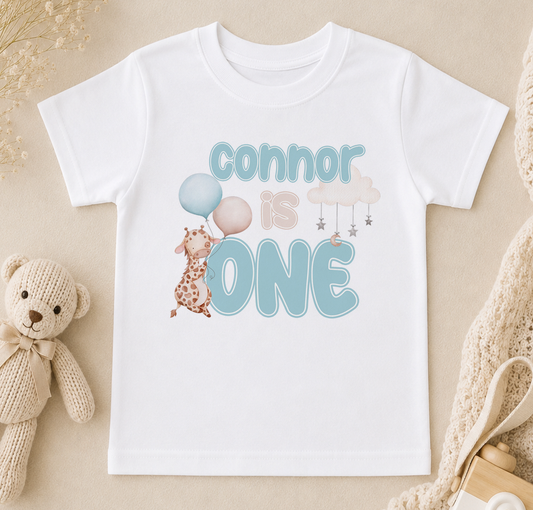 A white t-shirt with the text "Connor is one" printed on it, along with a giraffe and balloons. The t-shirt is placed on a beige surface, and there is a stuffed teddy bear and a beige blanket nearby.