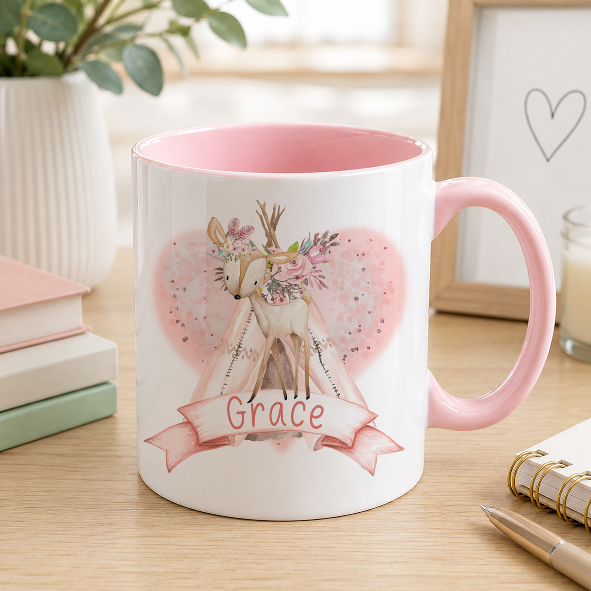 A pink and white mug with a design featuring a deer and flowers, along with the name "Grace" written on it. The mug is placed on a wooden surface, surrounded by various items such as books, a pen, and a candle.