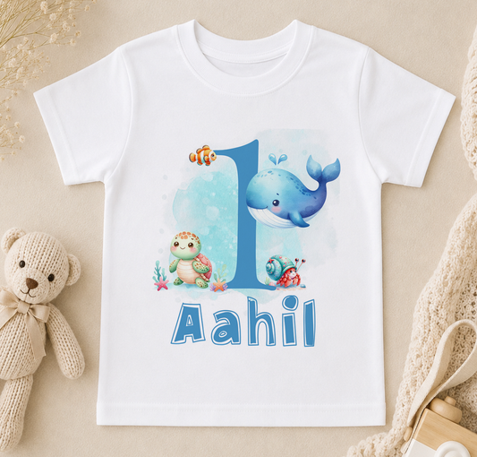 A white t-shirt with a blue whale and other sea creatures printed on it, along with the text "Aahil" below.