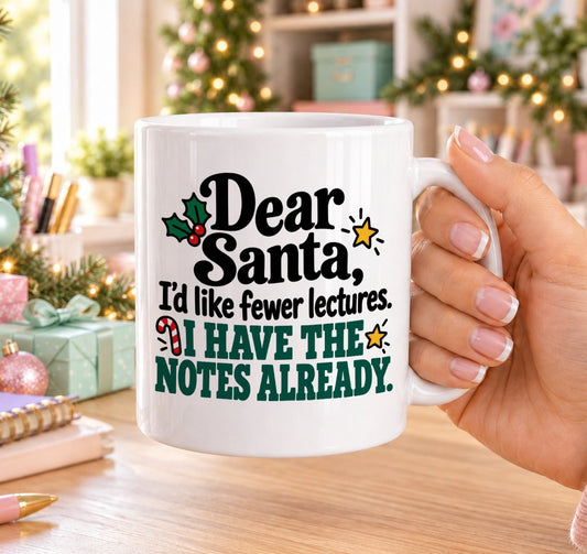 White 11oz ceramic Christmas mug printed with “Dear Santa, I’d like fewer lectures. I have the notes already.” in black and green lettering with holly, a candy cane and gold stars, held in a festive room