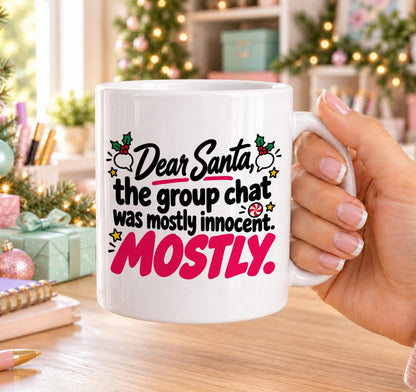 White 11oz ceramic Christmas mug printed with “Dear Santa, the group chat was mostly innocent. Mostly.” in black and pink lettering with holly, speech bubbles and gold stars, held in a festive room