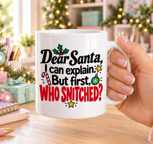 White 11oz ceramic Christmas mug printed with “Dear Santa, I can explain. But first, who snitched?” in black and red lettering with holly, a candy cane, a bauble and gold stars, held in a festive room