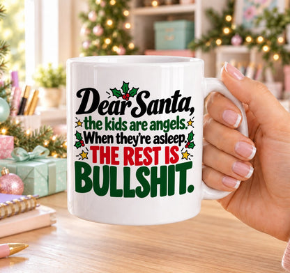White 11oz ceramic Christmas mug printed with “Dear Santa, the kids are angels. When they’re asleep. The rest is bullshit.” in red, green and black lettering with holly and gold stars, held in a festive room
