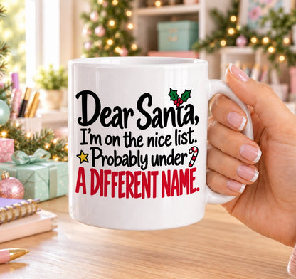 White 11oz ceramic Christmas mug printed with “Dear Santa, I’m on the nice list. Probably under a different name.” in black and red lettering with holly, a candy cane and gold stars, held in a festive room
