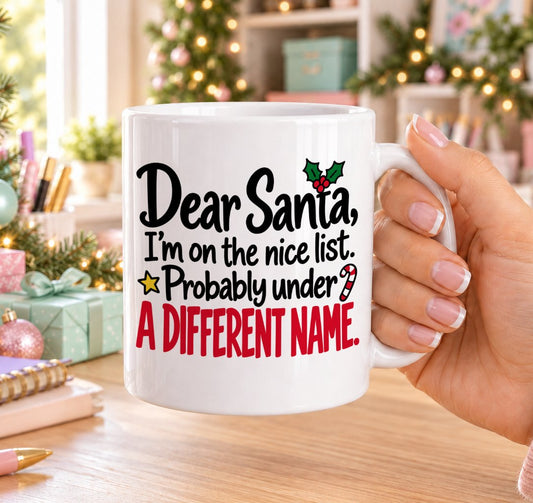 White 11oz ceramic Christmas mug printed with “Dear Santa, I’m on the nice list. Probably under a different name.” in black and red lettering with holly, a candy cane and gold stars, held in a festive room