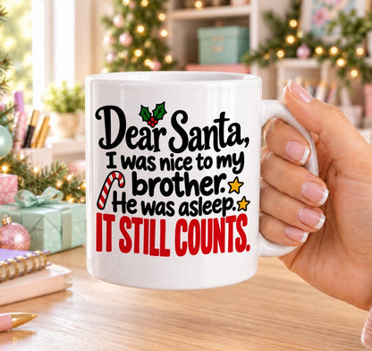 White 11oz ceramic Christmas mug printed with “Dear Santa, I was nice to my brother. He was asleep. It still counts.” in black and red lettering with holly, a candy cane and gold stars, held in a festive room