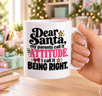 White 11oz ceramic Christmas mug printed with “Dear Santa, my parents call it attitude. I call it being right.” in black and pink lettering with a red bow and gold stars, held in a festive room