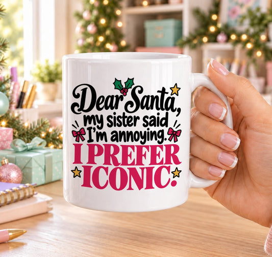White 11oz ceramic Christmas mug printed with “Dear Santa, my sister said I’m annoying. I prefer iconic!” in black and pink lettering with holly, pink bows and gold stars, held in a festive room