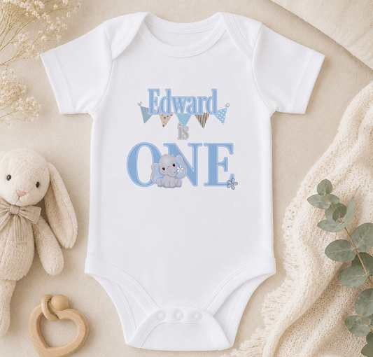 A white onesie with the text "Edward is one" printed on it, along with an image of a baby elephant. The onesie is placed on a beige surface, surrounded by a stuffed animal, a wooden toy, and some greenery.