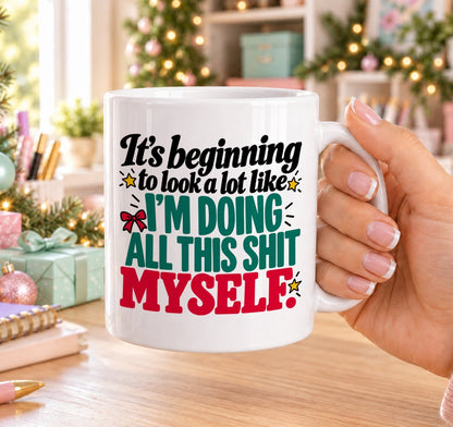 White 11oz ceramic Christmas mug printed with “It’s beginning to look a lot like I’m doing all this shit myself.” in red, green and black lettering with a red bow and gold stars, held in a festive room