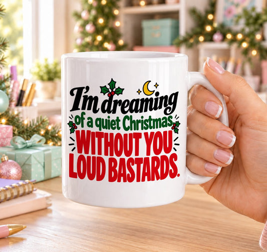 White 11oz ceramic Christmas mug printed with “I’m dreaming of a quiet Christmas without you loud bastards.” in red, green and black lettering with a crescent moon, stars and holly, held in a festive room