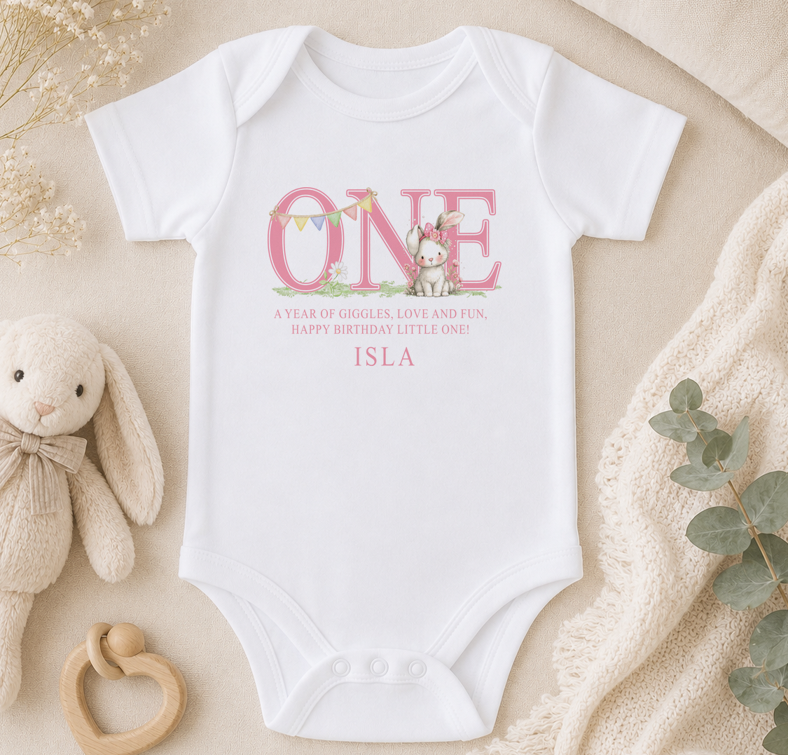Personalised white baby vest printed with a pink bunny, 'ONE' and a birthday message, styled on a bed with a soft toy bunny and a blanket.