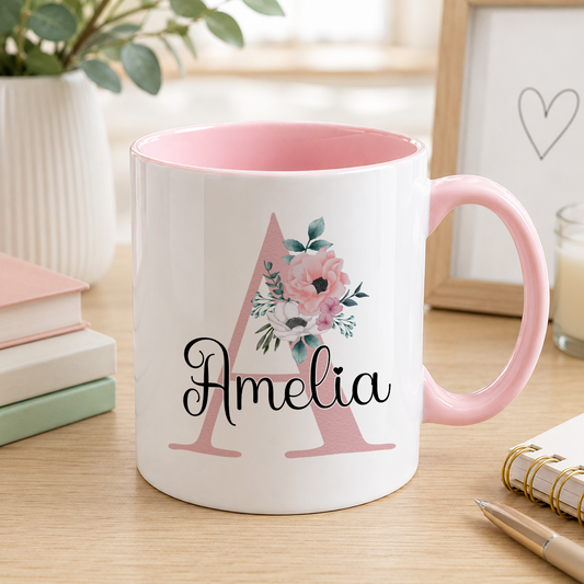 A pink and white mug with the name "Amelia" printed on it, sitting on a wooden surface next to some books and a plant.