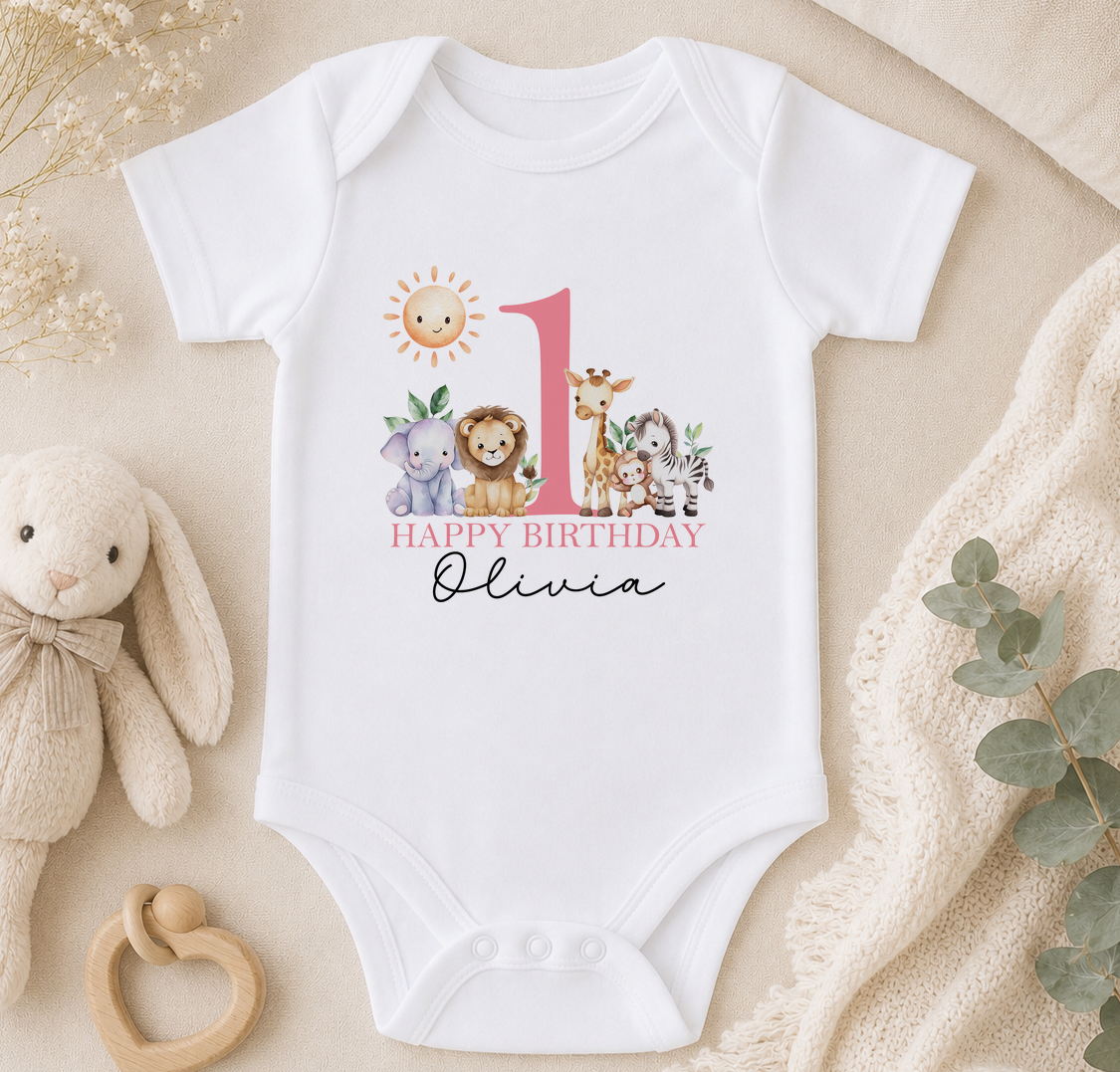 A white onesie with a pink number 1 and a happy birthday message for a child named Olivia, surrounded by stuffed animals and a beige blanket.