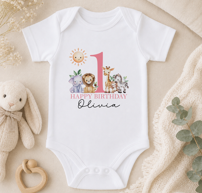 A white onesie with a pink number 1 and a happy birthday message for a child named Olivia, surrounded by stuffed animals and a beige blanket.