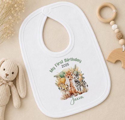 A white baby bib with a printed design of a giraffe, zebra, and other animals, along with the text "My First Birthday" and the year "2025". The bib is placed on a beige surface, with a stuffed animal toy and a wooden bead ring nearby.