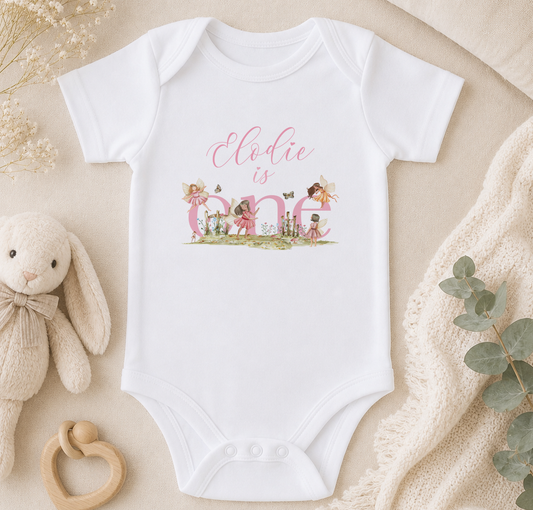 A white onesie with the text "Elodie is one" printed on it, surrounded by various baby items such as a stuffed bunny, a teething ring, and a blanket.