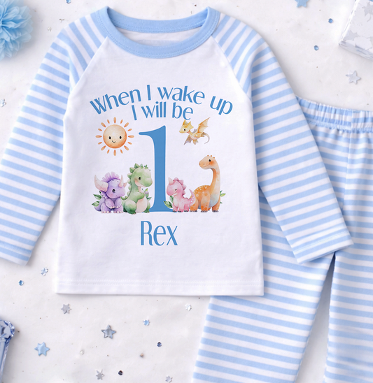 A white and blue pajama set with a dinosaur print and a personalized message for a first birthday.