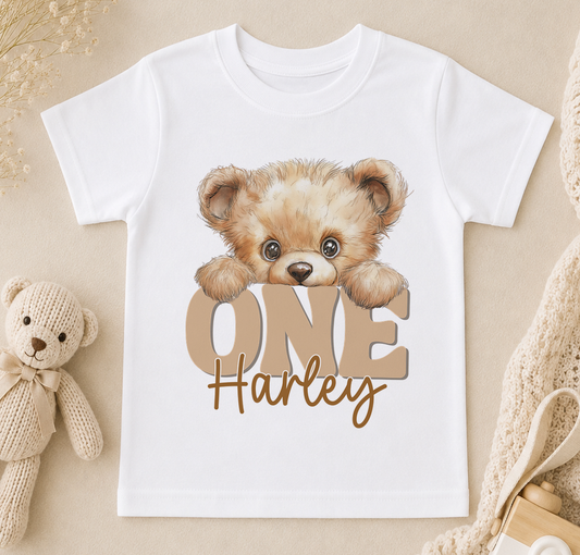 White t-shirt printed with a teddy bear design, the word 'ONE' and a personalised name, styled with a small stuffed bear.