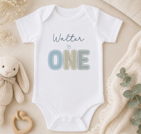 A white onesie with the text "Walter is one" printed on it, along with a wooden bib and a stuffed animal.