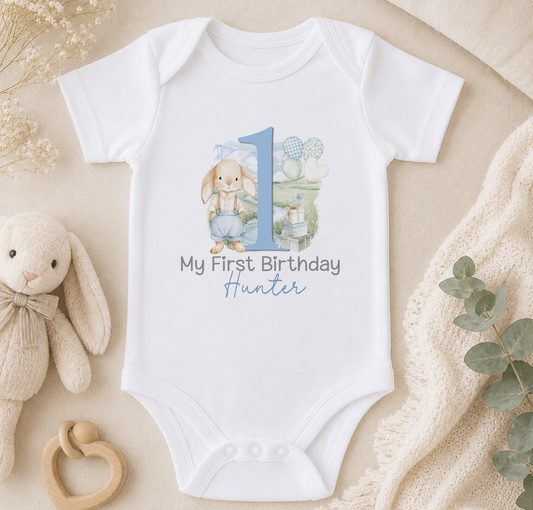 White baby onesie printed with a bunny in blue dungarees beside a large blue number '1', personalised with the sample name 'Hunter', styled on a bed with stuffed animals and a blanket.